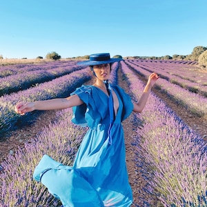 May include: A woman wearing a blue dress and a wide-brimmed blue hat stands in a field of lavender. The lavender is in rows and the woman is smiling.