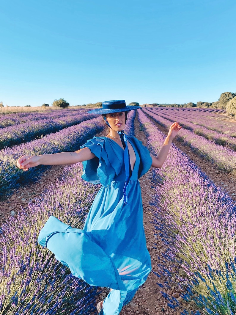 May include: A woman wearing a blue dress and a wide-brimmed blue hat stands in a field of lavender. The lavender is in rows and the woman is smiling.