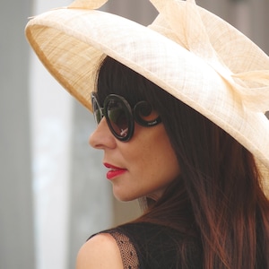 May include: A woman wearing a wide-brimmed, beige sun hat with a decorative bow. She is also wearing large, black sunglasses with the brand name "Prada" visible on the temple. Her dark hair and red lipstick provide contrast to the hat.