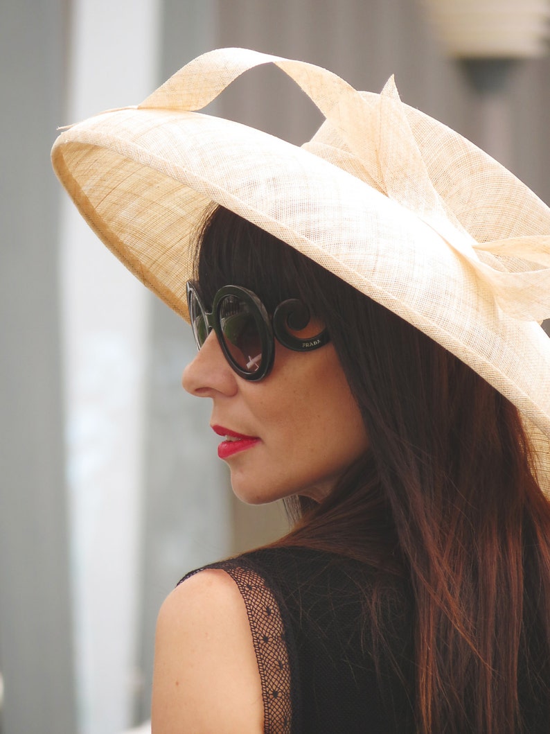 May include: A woman wearing a wide-brimmed, beige sun hat with a decorative bow. She is also wearing large, black sunglasses with the brand name "Prada" visible on the temple. Her dark hair and red lipstick provide contrast to the hat.