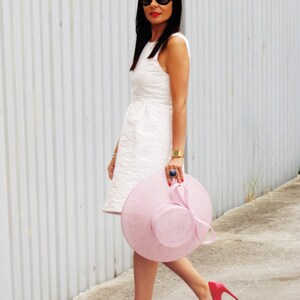 May include: A woman in a white dress, black sunglasses, and red heels walks along a sidewalk. She carries a large, light pink hat with a matching ribbon. A gold bracelet and a blue ring complete the look.
