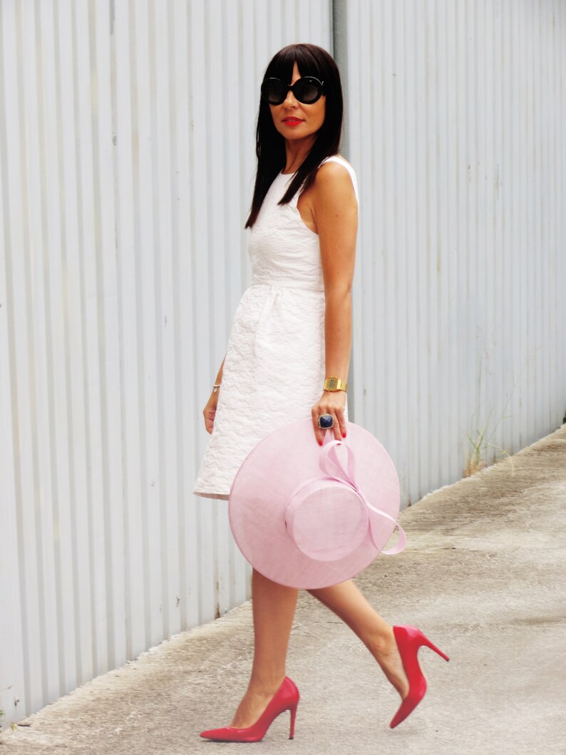May include: A woman in a white dress, black sunglasses, and red heels walks along a sidewalk. She carries a large, light pink hat with a matching ribbon. A gold bracelet and a blue ring complete the look.