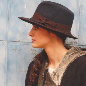 Op de afbeelding: Een vrouw draagt een donkerblauwe fedora met een bruin lint, een zwart jack met een bruine bontvoering en een lange vlecht.