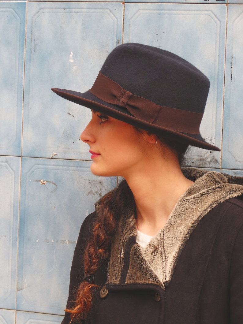 Op de afbeelding: Een vrouw draagt een donkerblauwe fedora met een bruin lint, een zwart jack met een bruine bontvoering en een lange vlecht.