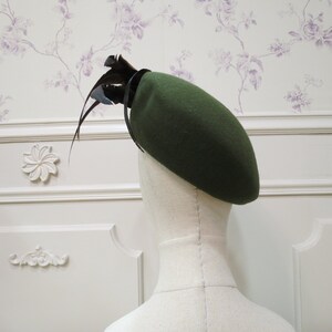 Op de afbeelding: Een donkergroene baret met een zwarte verenversiering. De baret is gemaakt van vilt en heeft een klassieke stijl.