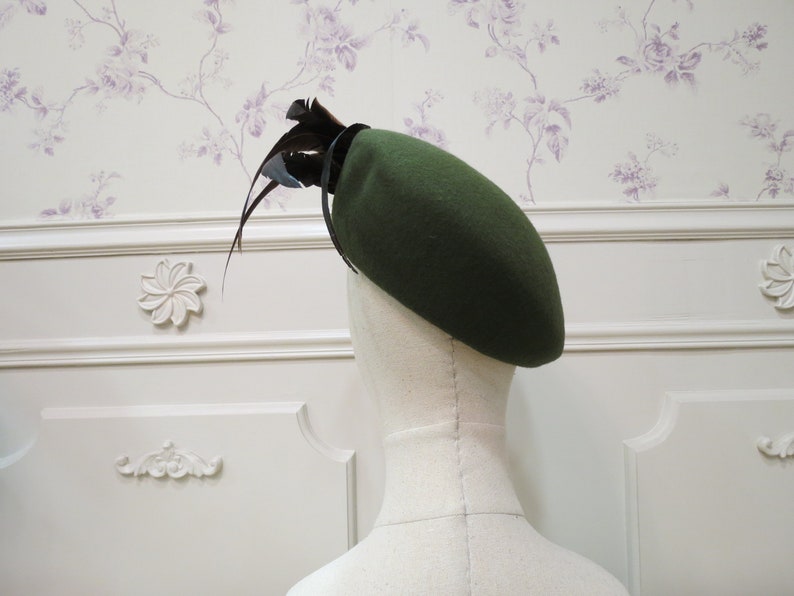 Op de afbeelding: Een donkergroene baret met een zwarte verenversiering. De baret is gemaakt van vilt en heeft een klassieke stijl.