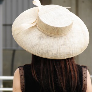 May include: A wide-brimmed, beige sun hat with a rounded crown and a decorative bow. The hat is made of woven material and is worn by a person wearing a black dress.