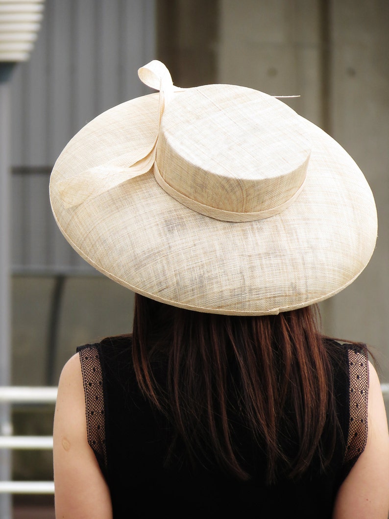 May include: A wide-brimmed, beige sun hat with a rounded crown and a decorative bow. The hat is made of woven material and is worn by a person wearing a black dress.