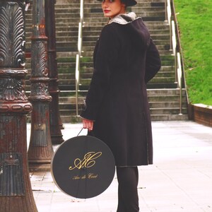 Op de afbeelding: Een vrouw in een zwarte jas en een zwarte fedora-hoed houdt een ronde zwarte doos vast met gouden letters die "An de C&oelig;ur" lezen.