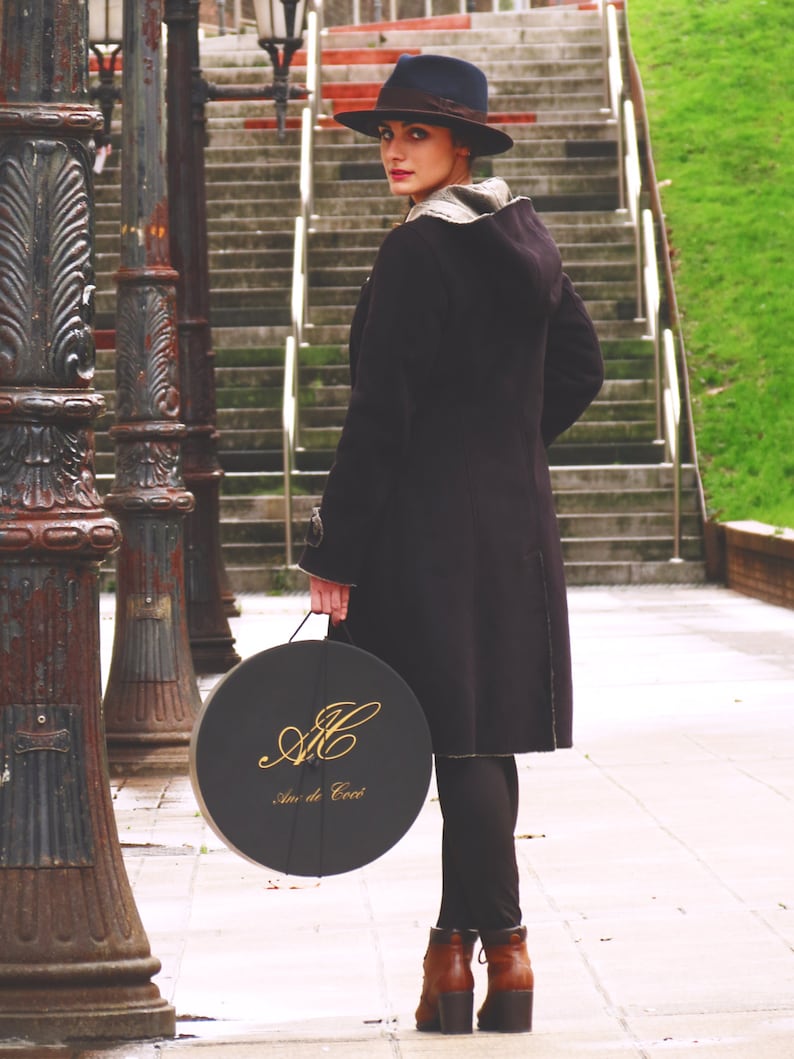 Op de afbeelding: Een vrouw in een zwarte jas en een zwarte fedora-hoed houdt een ronde zwarte doos vast met gouden letters die "An de C&oelig;ur" lezen.