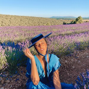 May include: A person wearing a blue dress and a blue hat sits in a field of lavender flowers. The person is looking at the camera and smiling.