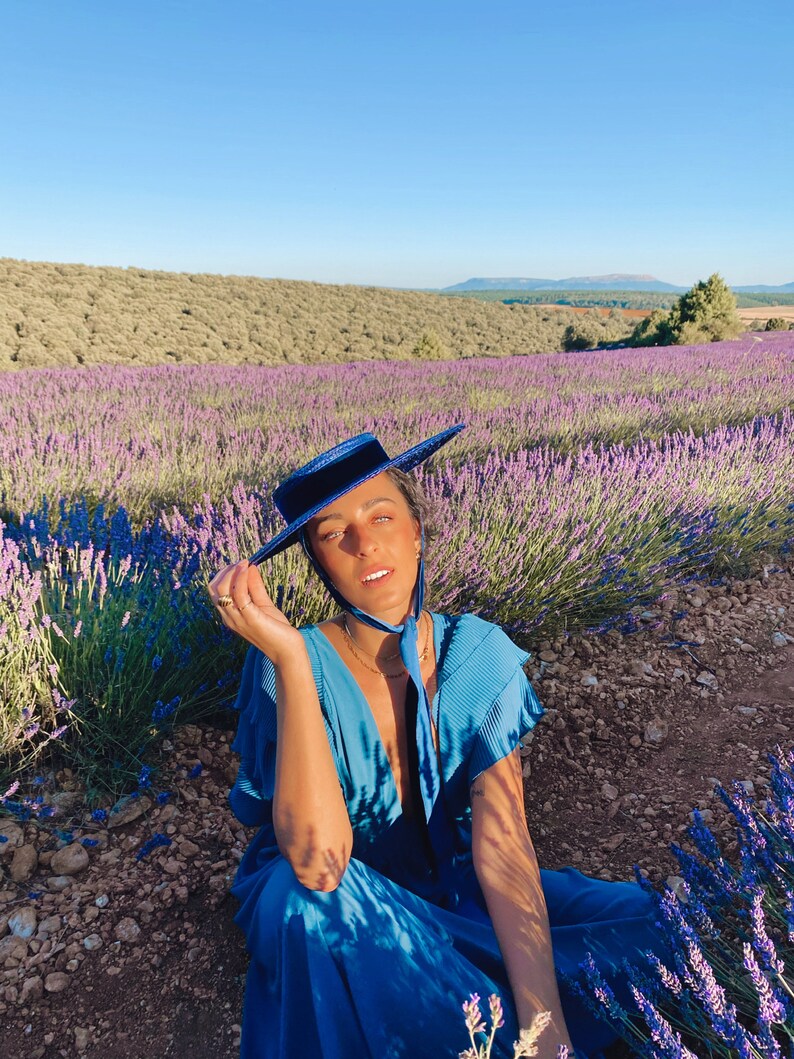 May include: A person wearing a blue dress and a blue hat sits in a field of lavender flowers. The person is looking at the camera and smiling.