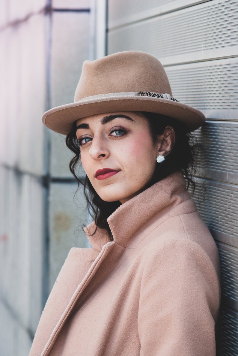 Op de afbeelding: Een vrouw draagt een bruine fedora met een verenversiering, een lichtbruine jas en een kleine zilveren oorbel. Ze kijkt met een serieuze uitdrukking naar de camera.