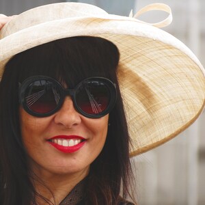 May include: A woman wearing a wide-brimmed beige hat and large black sunglasses with red-tinted lenses. The woman is smiling, with bright red lipstick. The hat has a cream-colored ribbon.