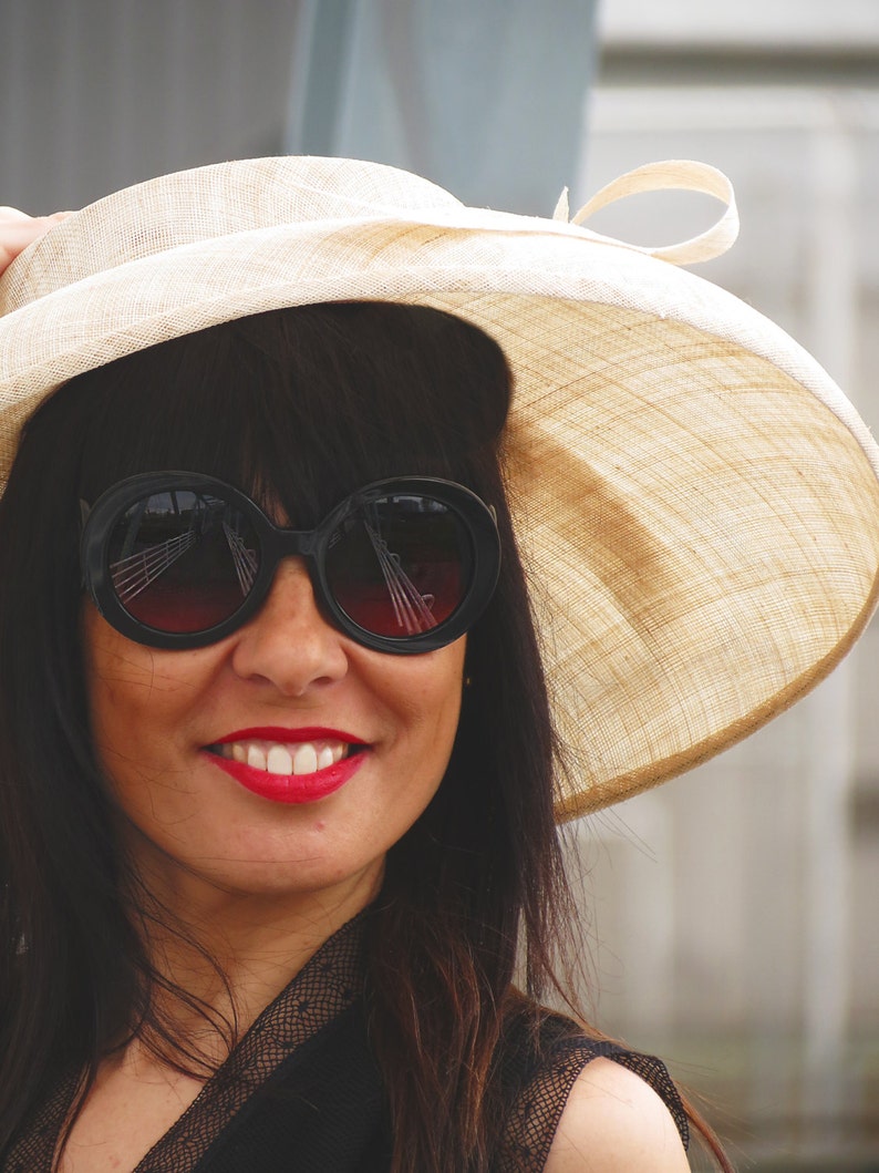 May include: A woman wearing a wide-brimmed beige hat and large black sunglasses with red-tinted lenses. The woman is smiling, with bright red lipstick. The hat has a cream-colored ribbon.