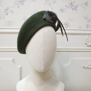 Op de afbeelding: Een donkergroene baret met een verenaccent. De baret is gemaakt van vilt en heeft een eenvoudig, klassiek ontwerp.