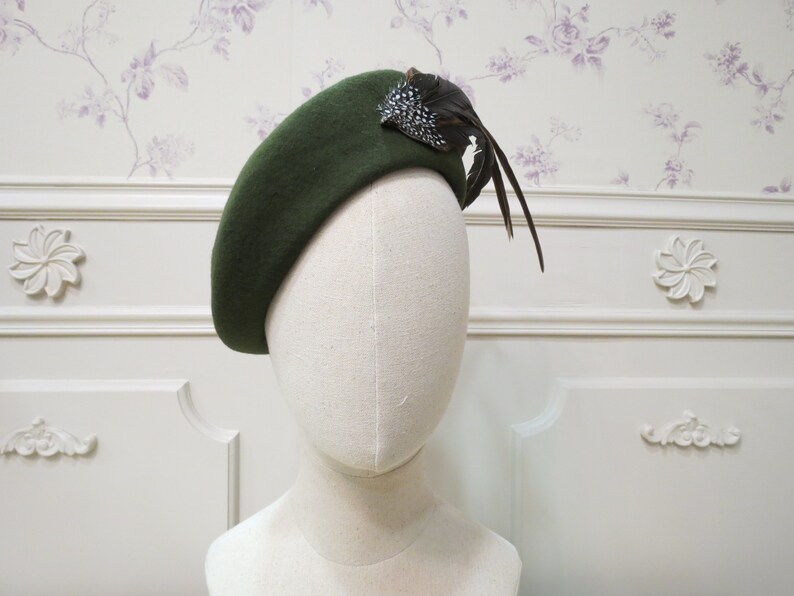 Op de afbeelding: Een donkergroene baret met een verenaccent. De baret is gemaakt van vilt en heeft een eenvoudig, klassiek ontwerp.