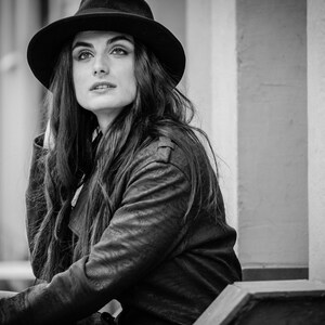 Op de afbeelding: Een vrouw draagt een zwarte fedora-hoed en een zwart leren jack. Ze zit op een trede met een muur achter haar.