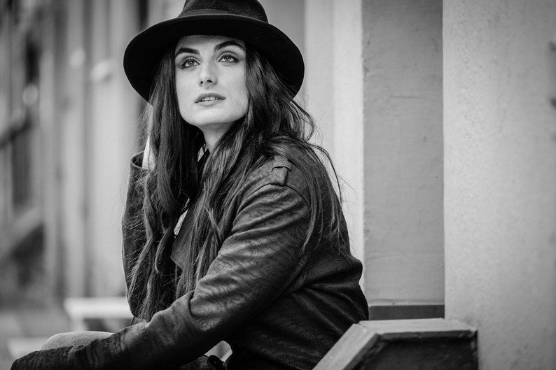 Op de afbeelding: Een vrouw draagt een zwarte fedora-hoed en een zwart leren jack. Ze zit op een trede met een muur achter haar.