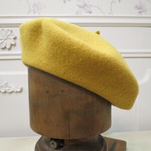 May include: A mustard yellow beret made of felt, displayed on a wooden mannequin head.