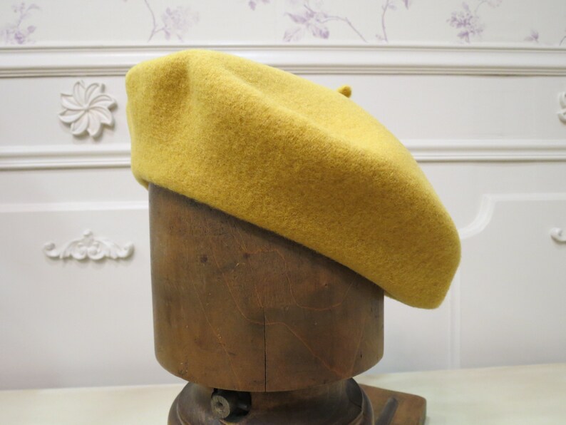 May include: A mustard yellow beret made of felt, displayed on a wooden mannequin head.