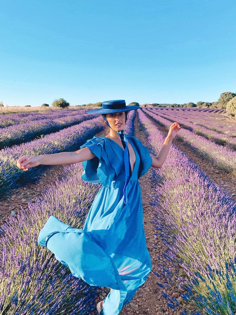 May include: A woman wearing a blue dress and a wide-brimmed hat stands in a field of lavender.