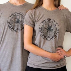 Könnte beinhalten: Zwei Personen tragen graue T-Shirts mit einem schwarzen Kreis-Motiv, auf dem "Respect Understanding" steht und eine stilisierte schwarze Blume im Kreis.