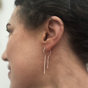 May include: Silver hoop earrings with a long, thin, straight bar hanging from the bottom of the hoop.