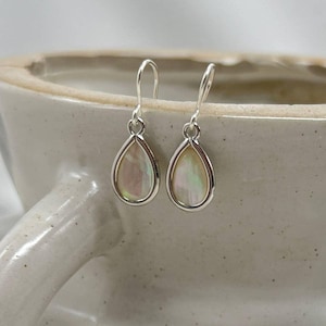 May include: A pair of teardrop-shaped earrings with iridescent, pearlescent centers and silver-colored metal frames. The earrings hang from curved hooks. The background is a neutral-toned ceramic container.