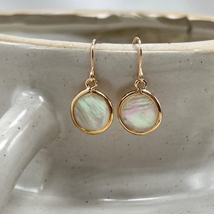May include: A pair of gold-coloured drop earrings. Each earring features a round, iridescent, mother-of-pearl disc set within a gold-tone bezel. The earrings hang from a simple gold-coloured hook.