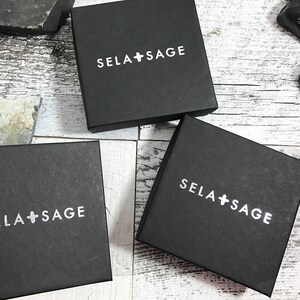 May include: Three square black gift boxes with the white text SELA + SAGE printed on the top lid of each box. The boxes are arranged in a cluster, each featuring a clean, matte finish and a simple, bold sans-serif font for the brand name.