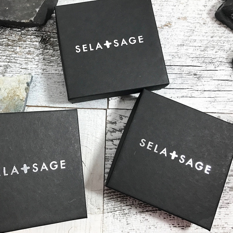 May include: Three square black gift boxes with the white text SELA + SAGE printed on the top lid of each box. The boxes are arranged in a cluster, each featuring a clean, matte finish and a simple, bold sans-serif font for the brand name.