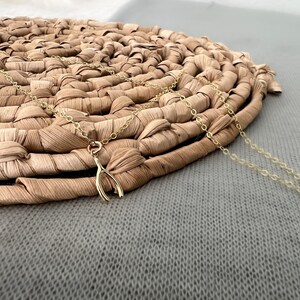 May include: A gold necklace with a small, delicate wishbone charm. The necklace is laying on a woven straw mat.