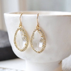 May include: A pair of gold dangle earrings with clear crystal stones set in a gold halo. The earrings have a teardrop shape.