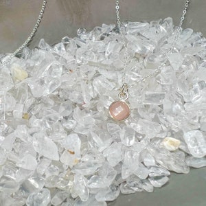 Puede incluir: Un collar de cadena de plata con un colgante de piedra rosa. El collar est&aacute; sobre un lecho de cristales de cuarzo blanco.