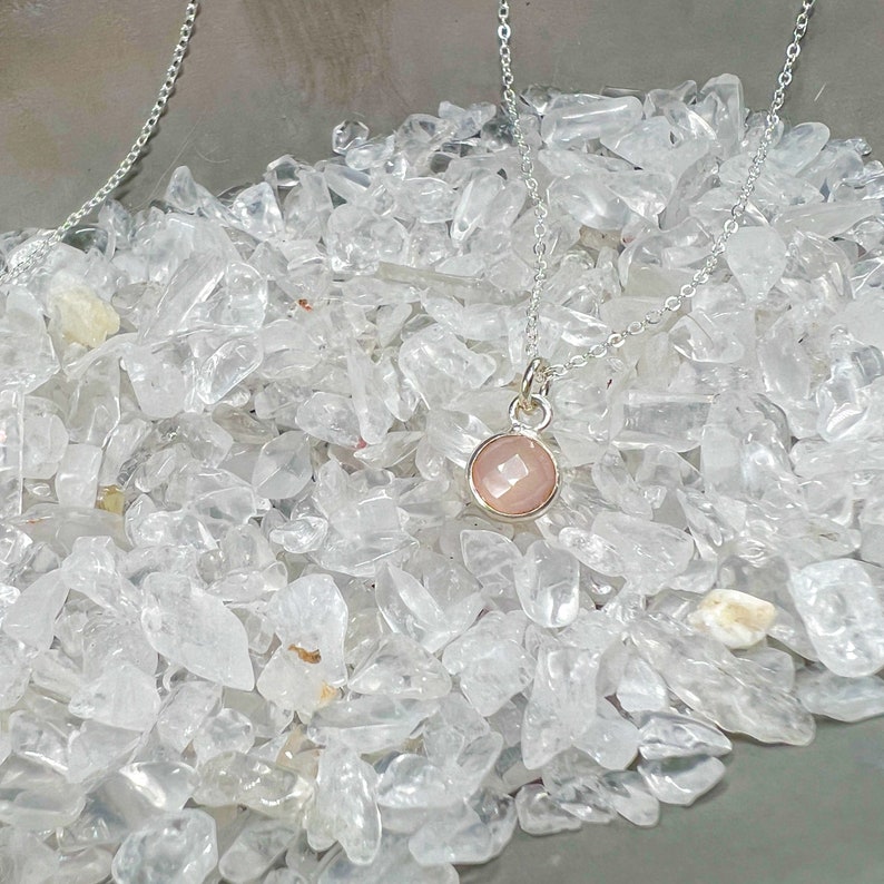 Puede incluir: Un collar de cadena de plata con un colgante de piedra rosa. El collar est&aacute; sobre un lecho de cristales de cuarzo blanco.