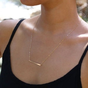Minimalist gold bar necklace thin horizontal bar design, dainty gold filled layering necklace, simple modern everyday jewelry.   A gold bar necklace with a delicate chain. The necklace is worn by a person with a black tank top.