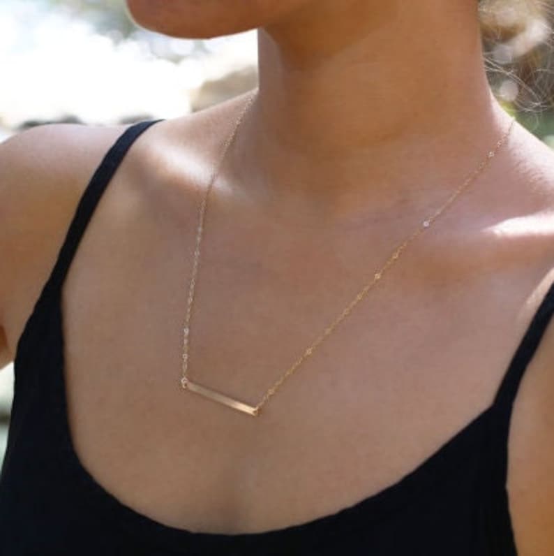 Minimalist gold bar necklace thin horizontal bar design, dainty gold filled layering necklace, simple modern everyday jewelry.   A gold bar necklace with a delicate chain. The necklace is worn by a person with a black tank top.