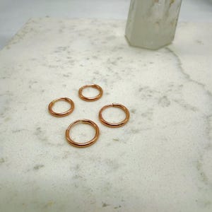 May include: Four small, round, rose gold hoop earrings are arranged on a white marble surface. The earrings are evenly spaced and appear to be made of a shiny metal. A clear, faceted crystal is in the background.