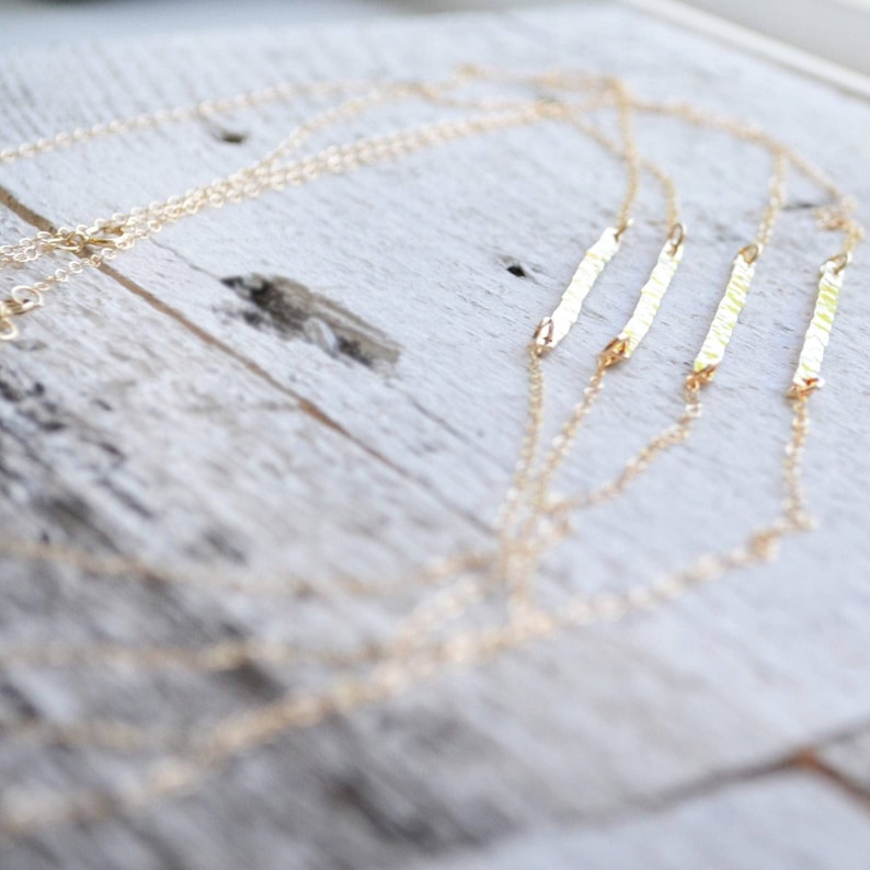 A gold filled necklace with four thin, hammered bar pendants. The necklace is layered on a white wooden surface. Minimalist gold bar necklace with modern dainty jewelry aesthetic, clean simple look.