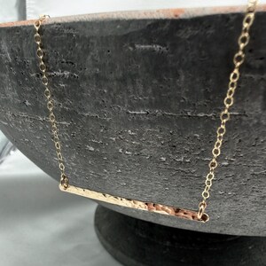 May include: A gold-toned necklace featuring a thin, horizontal bar pendant with a hammered, textured surface. The pendant is suspended from a delicate, fine-link chain that connects to each end of the bar, creating a simple, clean design.