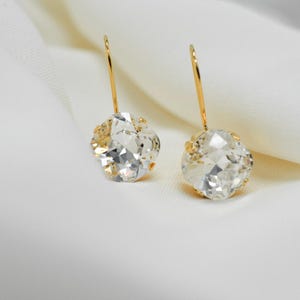 Puede incluir: Un par de pendientes de aro de oro con piedras de cristal transparentes. Las piedras son redondas y tienen un diseño facetado.