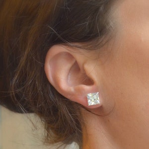 May include: Close-up of a square-cut, clear gemstone stud earring on a person's ear. The earring is set in a silver or white metal setting. The person has brown hair.