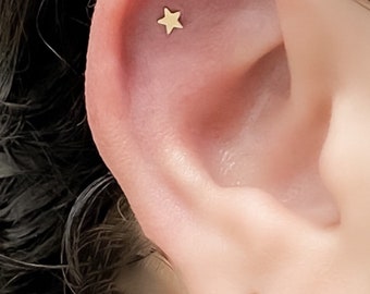 Puede incluir: Primer plano de la oreja de una persona con un pequeño pendiente de estrella de oro. El pendiente está en el lóbulo superior de la oreja.