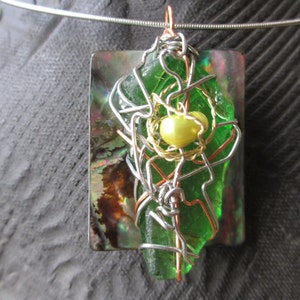 May include: A green glass pendant encased in a wire cage with a yellow pearl in the center. The pendant is set against a background of iridescent abalone shell.
