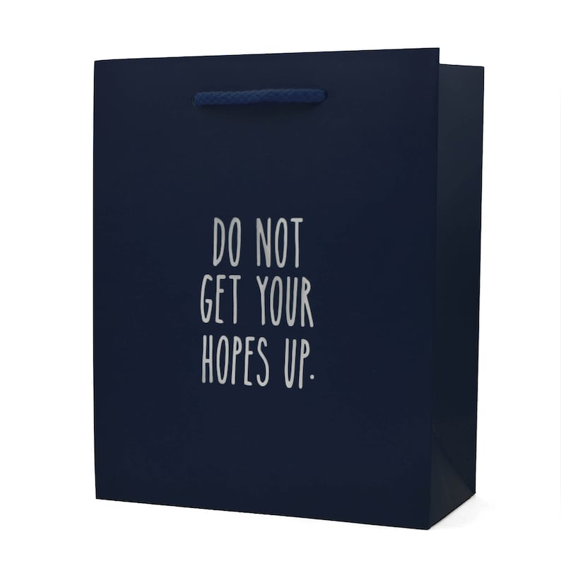 Puede incluir: Una bolsa de regalo azul oscuro con un asa de cuerda blanca. La bolsa tiene el texto "DO NOT GET YOUR HOPES UP." impreso en la parte delantera.