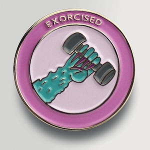 May include: A pink and gold enamel pin with a green monster hand holding a gray dumbbell. The pin says "EXORCISED" in gold letters.