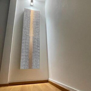 a hallway with a wooden floor and white walls. There is a large piece of artwork hanging on the wall, and a light fixture is visible above it.