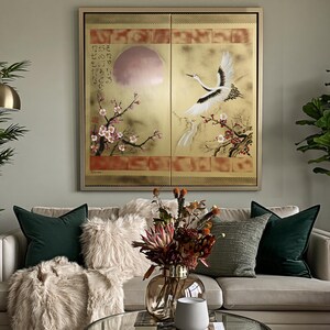 a living room with a large, ornate painting of a crane and cherry blossoms hanging on the wall above a beige sofa. The sofa is adorned with green and gray throw pillows, and a vase of flowers is placed on the coffee table in front of the sofa.