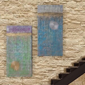 two abstract paintings hanging on a brick wall, with a staircase visible in the background.